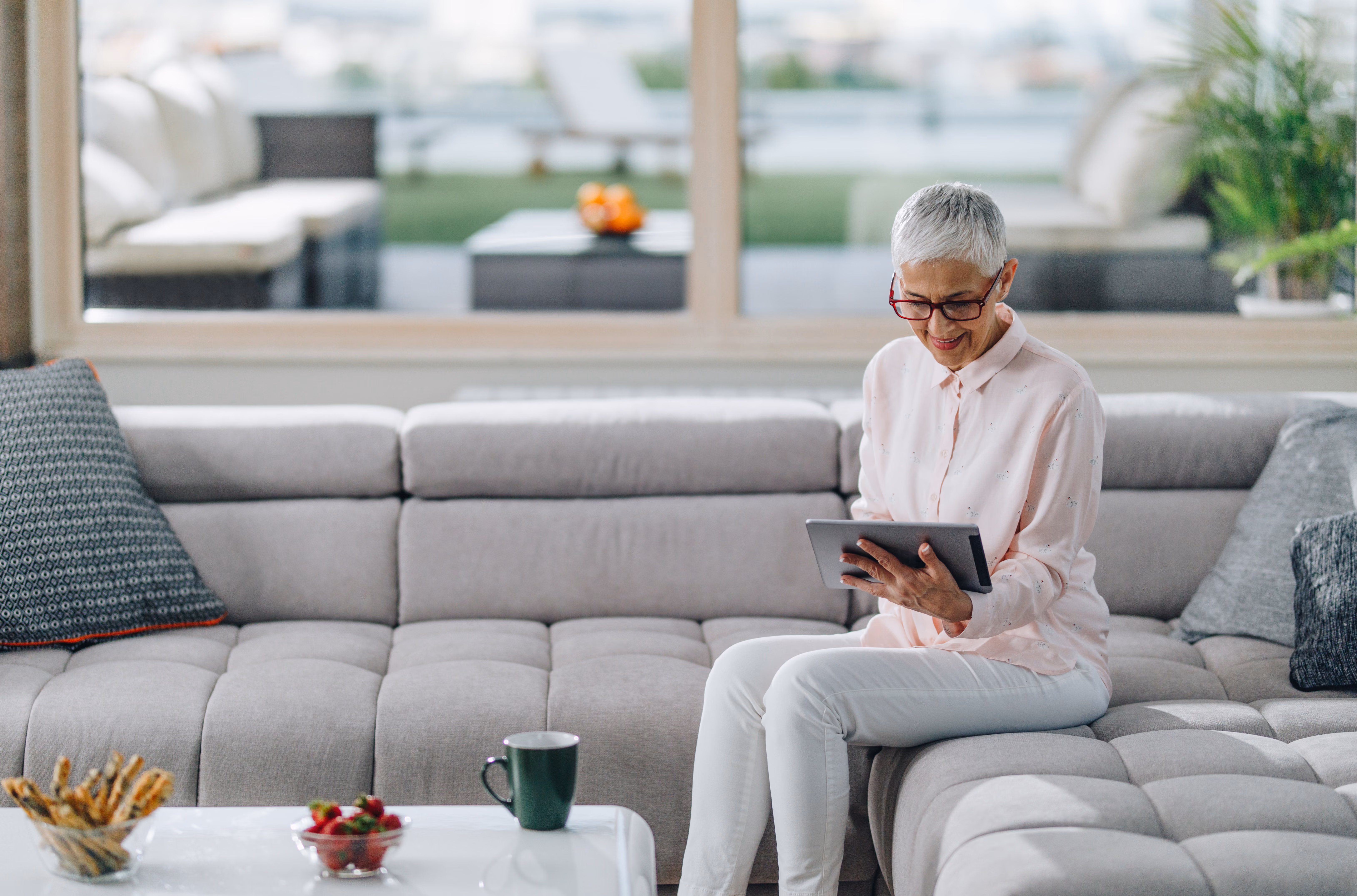 A woman with white hair checks her tablet while seated on a gray couch in a contemporary home.