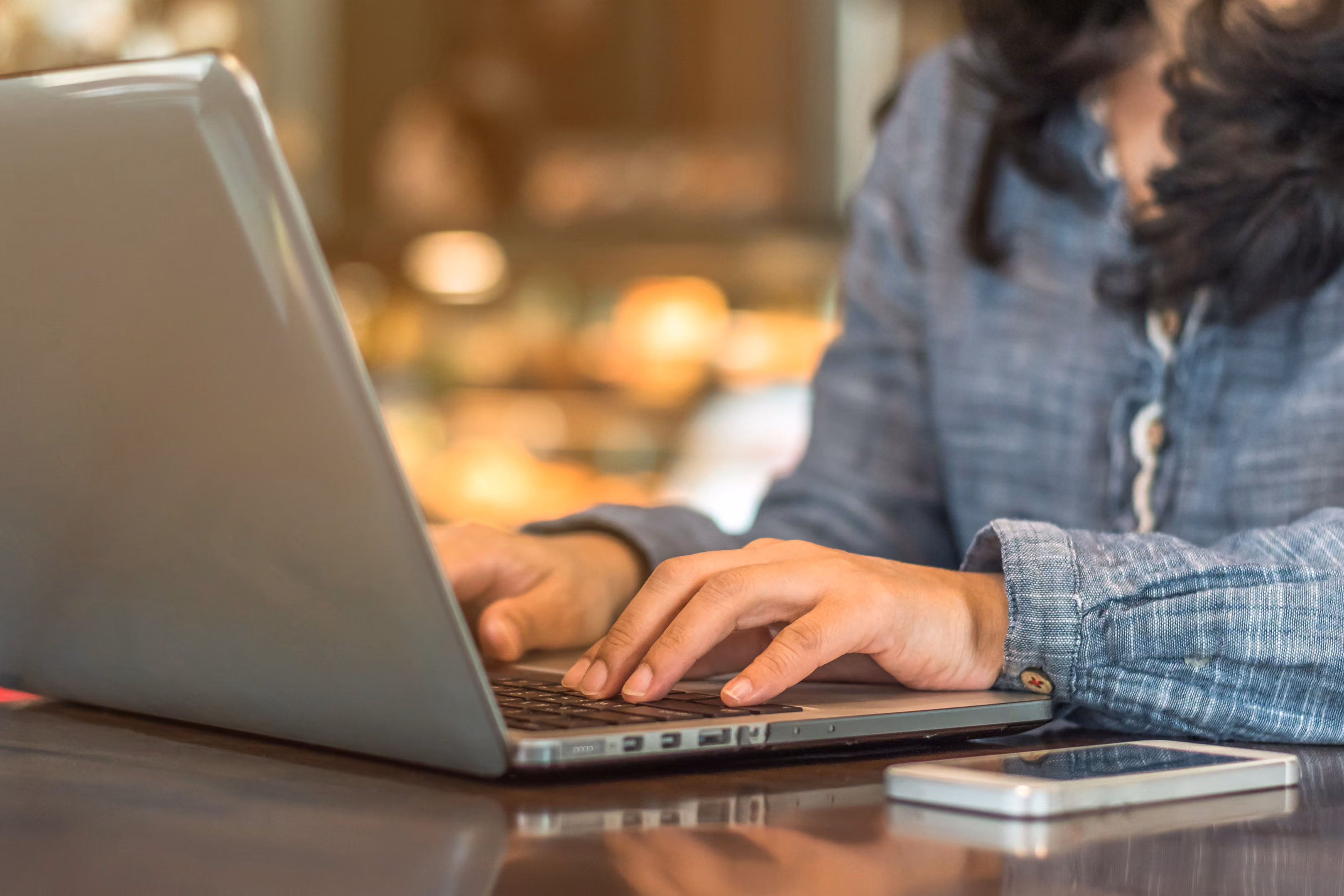 Close crop of a woman typing on her laptop, with her smartphone on the table next to her