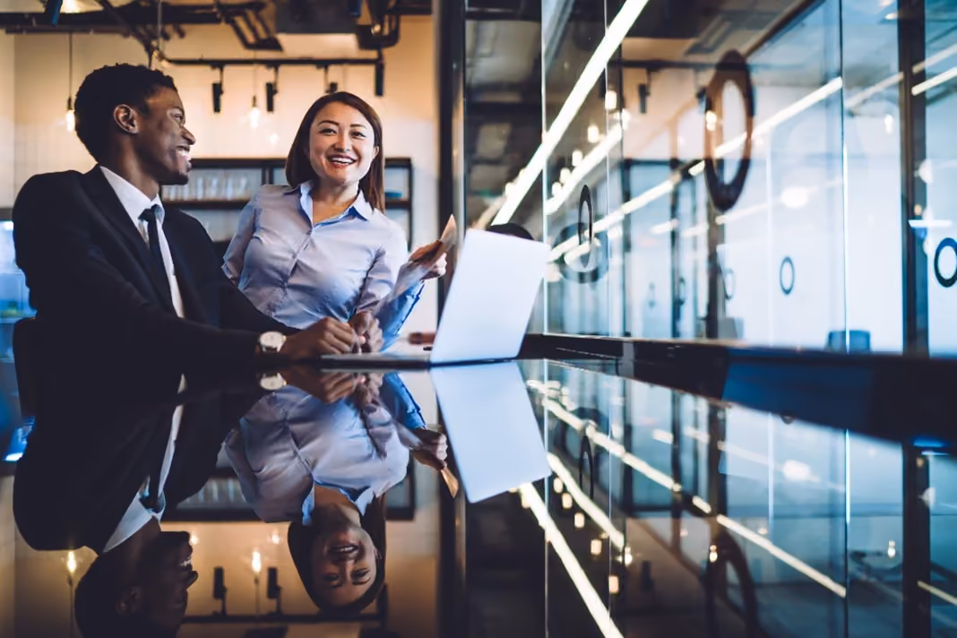 Two young professionals are laughing and smiling in the office after seeing positive changes and results on their laptop.